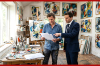 An artist in paint-stained clothing and a lawyer in a suit discuss a contract in a bright art studio filled with abstract paintings